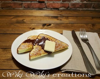 May include: Two slices of French toast on a white plate with chocolate sauce and a pat of butter. The plate is on a wooden table with a brown linen napkin and silver cutlery.
