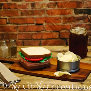 May include: A deli sandwich with ham, lettuce, and tomato on white bread sits on a wooden cutting board. A metal bowl of potato chips, a glass of iced tea, and salt and pepper shakers complete the meal. A fork and knife rest on a linen napkin.