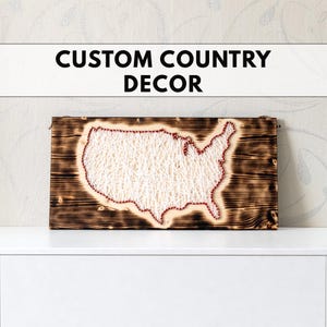 Könnte beinhalten: Ein Holzschild mit dem Text "CUSTOM COUNTRY DECOR" über einer String-Art-Karte der Vereinigten Staaten. Die Karte besteht aus weißem Faden und roten Nadeln, vor einem dunklen, verbrannten Holzgrund.