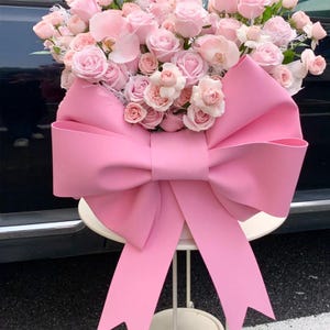 May include: A large arrangement of pink roses and orchids, overflowing from a white container. A large, bright pink bow is the focal point, adding a decorative touch to the floral display. The arrangement is set on a small, white table.