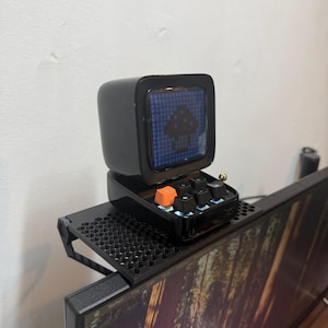 May include: A black retro digital display with a mushroom icon on the screen, and a small keyboard with an orange key. The device is mounted on a black shelf above a computer monitor.