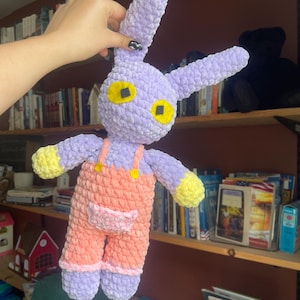 Puede incluir: Un juguete de conejo de ganchillo con cuerpo de color morado claro, orejas largas y manos amarillas. El conejo lleva un peto de color melocotón con un pequeño bolsillo rosa en la parte delantera y botones amarillos. Tiene dos grandes ojos amarillos con cuadrados negros.