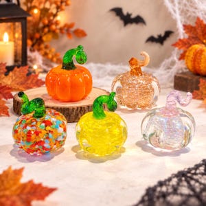 May include: Five decorative glass pumpkins with curled stems. One is solid orange, one is clear with multicolored confetti, one is bright yellow, one is clear with gold glitter, and one is clear with an iridescent finish, all arranged together.