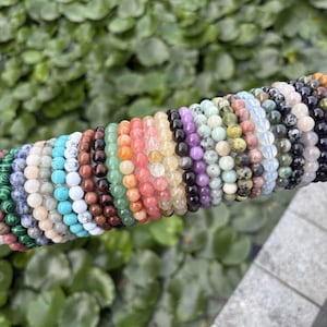 May include: A collection of beaded bracelets in various colors and stone types. The bracelets are displayed on a white cylindrical form, showcasing a range of hues including green, blue, pink, and brown. The beads appear to be approximately 0.3 inches in diameter.