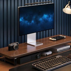 May include: A contemporary desk arrangement with a computer monitor displaying a starry sky, a wooden monitor stand, a camera, a keyboard, and a mouse. A black desk lamp and a games console are also present. The desk is made of wood.