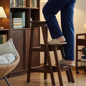 May include: A dark brown wooden step stool with three steps. The step stool is being used by someone wearing blue pants and beige slippers. The step stool is in front of a wooden bookcase.