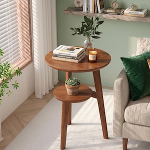 Pode incluir: Uma mesa lateral redonda de madeira de dois níveis com acabamento castanho quente. O nível superior contém livros e uma vela, enquanto o nível inferior exibe uma suculenta em vaso. A mesa tem 61 cm de altura.