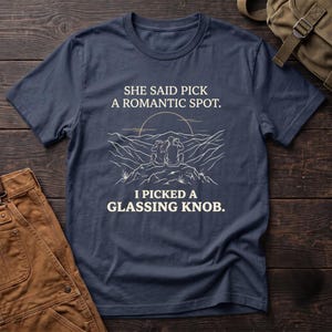 May include: A navy blue short-sleeve t-shirt featuring a white line art graphic of two people sitting on a mountain peak looking at a sunset. The text reads: SHE SAID PICK A ROMANTIC SPOT. I PICKED A GLASSING KNOB. The shirt has a crew neckline.