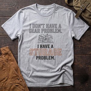 May include: A light gray short-sleeved t-shirt featuring the black text I DON'T HAVE A GEAR PROBLEM. I HAVE A STORAGE PROBLEM. with a central line drawing of various outdoor equipment and the word STORAGE printed in orange block letters.