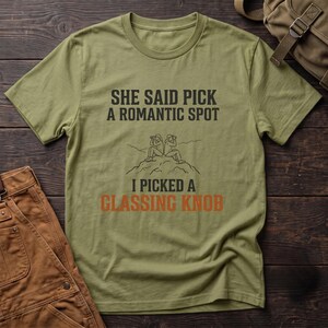 May include: An olive green crew neck t-shirt featuring black text that reads: SHE SAID PICK A ROMANTIC SPOT I PICKED A GLASSING KNOB. Below the first line of text is a black line drawing of two people sitting on a mountain peak.