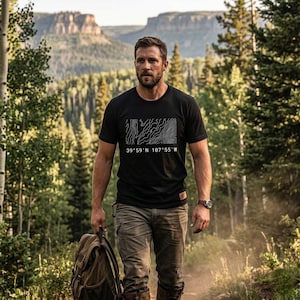 Puede incluir: Camiseta negra con un diseño topográfico blanco y coordenadas 39°59'N 107°55'W. La camiseta se usa con pantalones marrones y una mochila marrón. El fondo muestra un paisaje montañoso y árboles.