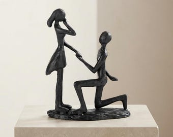 May include: A dark charcoal-colored sculpture depicting two abstract figures. One figure stands upright while the other kneels on one knee, reaching out to hold the hand of the standing figure. The piece has a textured, matte finish throughout.