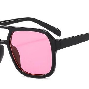 May include: Black square-framed sunglasses with pink lenses. The sunglasses have a double bridge and thick arms. The lenses are a vibrant pink colour, and the frames are a solid black.