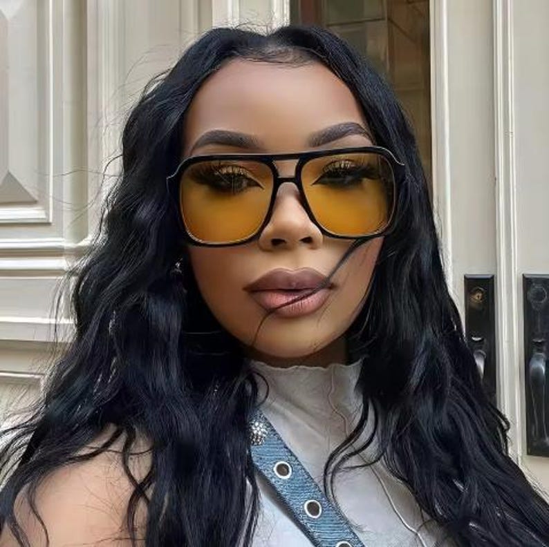 May include: Black-framed sunglasses with yellow-tinted lenses. The sunglasses have a square shape and a double bridge. The person in the image has long, dark hair and is wearing a white top.