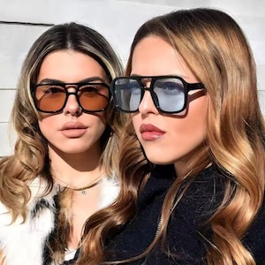 May include: Two people wearing black square sunglasses with tinted lenses. One pair has orange lenses, and the other has blue-grey lenses. The sunglasses have a thick black frame and a double bridge design.