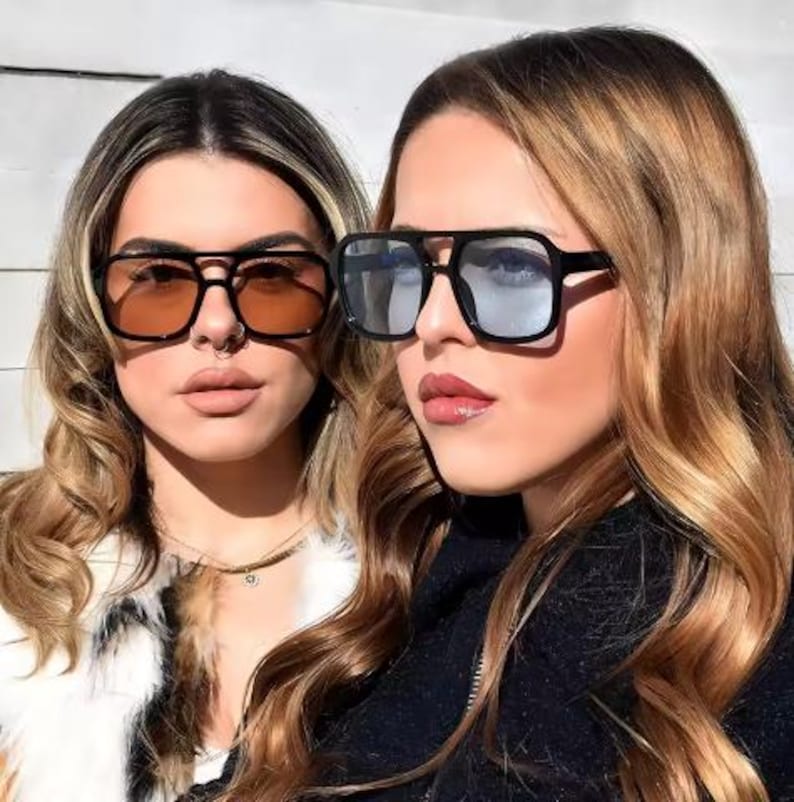 May include: Two people wearing black square sunglasses with tinted lenses. One pair has orange lenses, and the other has blue-grey lenses. The sunglasses have a thick black frame and a double bridge design.