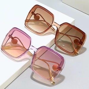 May include: Two pairs of oversized sunglasses with square frames. One pair has a gradient pink and orange lens, the other has a gradient brown lens. Both have gold-colored accents and a decorative circular element near the hinges.