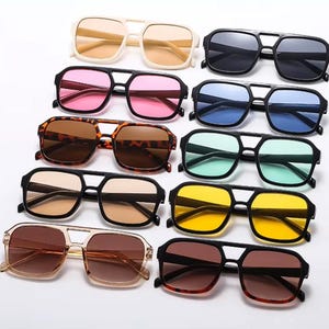 May include: An assortment of sunglasses with different coloured lenses, such as pink, blue, yellow, and brown. The frames are black, tortoiseshell, and cream. The sunglasses feature a double bridge design.