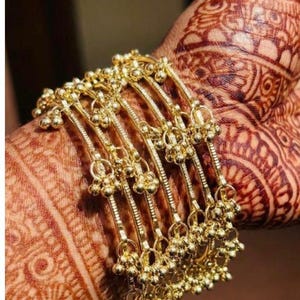 Puede incluir: Una colección de pulseras doradas, cada una adornada con pequeñas decoraciones esféricas. Las pulseras están apiladas en una muñeca decorada con intrincados diseños de henna, mostrando una estética tradicional.