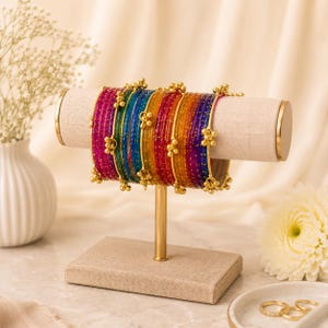 May include: A display of colorful bangles in shades of pink, green, blue, yellow, orange, and purple. The bangles are adorned with gold-colored accents and are arranged on a beige stand with gold trim. The stand is on a beige base.