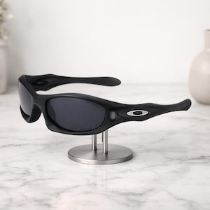 May include: Black sunglasses with dark lenses, presented on a silver stand. The sunglasses feature a curved, sporty design with a prominent frame and a silver logo on the temples. The stand has a circular base.