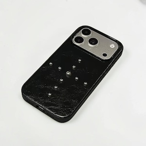 Puede incluir: Una funda de smartphone negra con una superficie texturizada similar al cuero, que presenta un patrón de cruz hecho de pequeños remaches metálicos plateados. La funda tiene un recorte rectangular en la parte superior para la cámara triple y el flash.