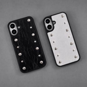 May include: Two rectangular phone cases with black edges and silver metal studs. One case features a textured black surface, while the other has a smooth white surface. Both cases include a circular cutout for a dual-lens camera system.