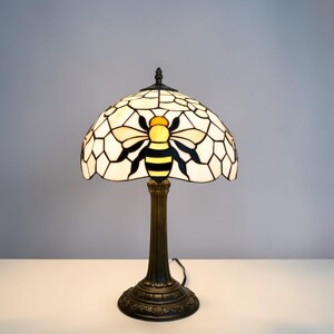 May include: A stained glass table lamp featuring a bee design. The lamp has a cream-colored shade with a geometric pattern and a yellow and black bee in the center. The base and stem are dark bronze-colored.