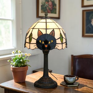 May include: A stained glass table lamp featuring a black cat design on the shade. The lamp has a bronze-colored base and a cream-colored shade with green and brown accents. The cat's eyes are yellow. The lamp is on a wooden table.