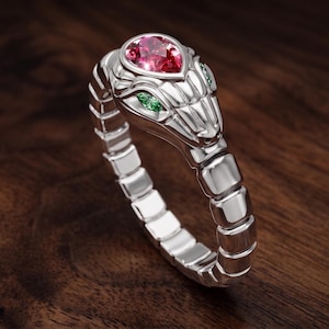 Pode incluir: Um anel prateado em forma de serpente enrolada com corpo segmentado. A cabeça da serpente apresenta uma pedra preciosa rosa em forma de pera no topo e duas pequenas pedras verdes como olhos, tudo montado numa banda metálica polida.