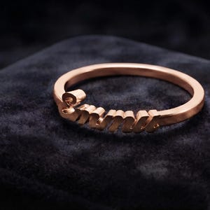 Pode incluir: Um anel em tom de ouro rosa com um design de placa de nome personalizado. A banda é fina e circular, com o nome "Emma" integrado na secção frontal numa fonte cursiva metálica. A superfície metálica tem um acabamento liso e polido.