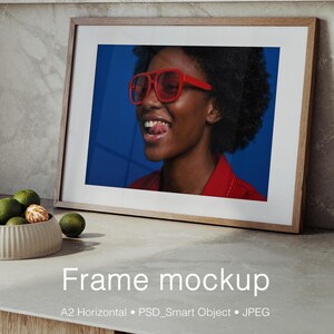 May include: A horizontal wooden picture frame containing a photograph of a person with dark curly hair wearing red-tinted aviator sunglasses and a red shirt against a solid blue background. The text Frame mockup A2 Horizontal PSD_Smart Object JPEG is visible.