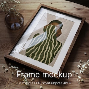 May include: A framed art print featuring a figure with green and white striped patterns. The artwork is in a dark wood frame with a white mat. The image is on a wooden surface with a glass vase and small white flowers.