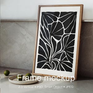 Pode incluir: Uma moldura de madeira retangular vertical contendo uma impressão branca com um padrão floral abstrato preto arrojado. A obra de arte apresenta formas orgânicas estilizadas semelhantes a folhas que preenchem a moldura.
