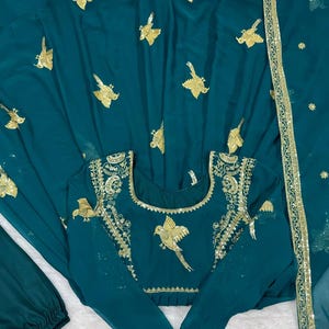 Puede incluir: Conjunto de ropa formal de color verde azulado con bordados dorados de pájaros. El conjunto incluye una parte superior de manga larga con detalles dorados alrededor del cuello y las mangas, pantalones a juego y una bufanda drapeada. La tela tiene un aspecto fluido.