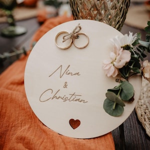 Könnte beinhalten: Ein runder Holz-Hochzeitsringhalter mit den eingravierten Namen "Nina & Christian". Zwei goldene Ringe sind mit Bindfaden gebunden. Der Halter ist mit Blumen und einem Herz-Ausschnitt verziert. Im Hintergrund ein Glas und orangefarbener Stoff.
