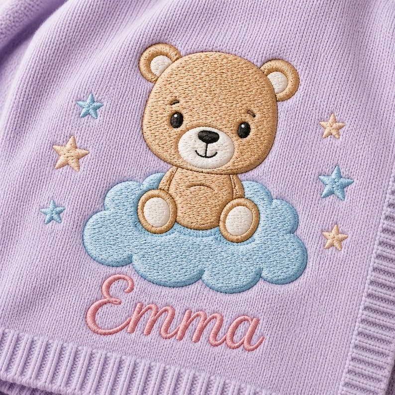 May include: A light purple knit fabric featuring an embroidered design of a brown teddy bear sitting on a light blue cloud. The bear is surrounded by small light blue and beige stars. The name Emma is embroidered in pink cursive script below.