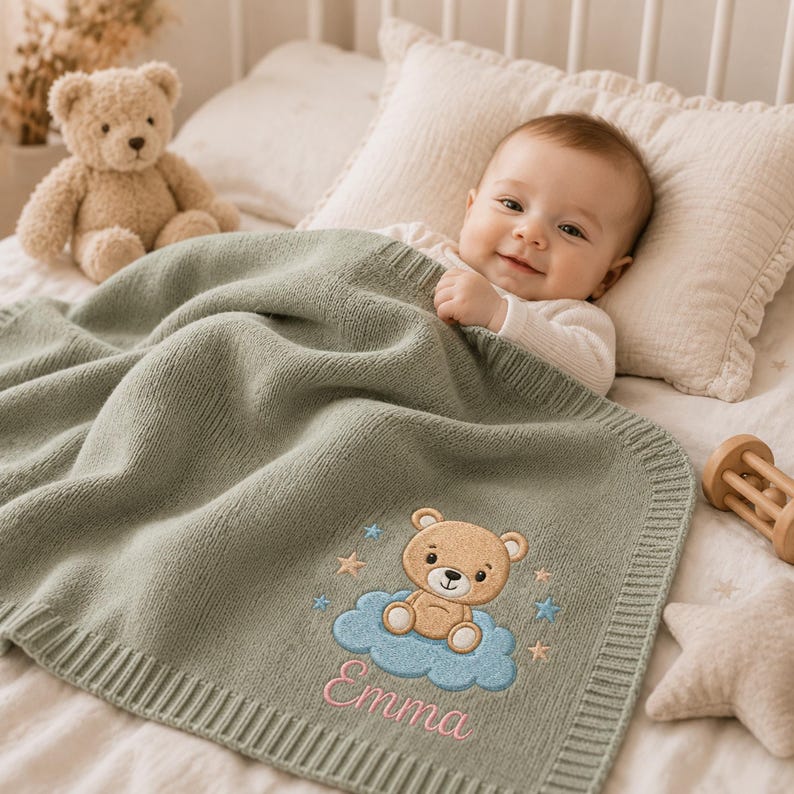 May include: A sage green knitted baby blanket featuring an embroidered design of a light brown teddy bear sitting on a blue cloud, surrounded by small stars. The name Emma is embroidered in pink script below the cloud, with a ribbed border.