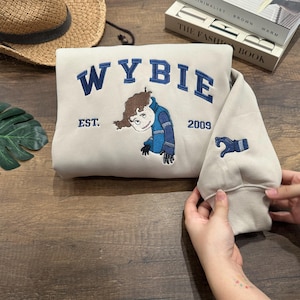 May include: Beige sweatshirt with the word "WYBIE" embroidered in navy blue, along with a cartoon character and the text "EST. 2009." The sleeve features a navy blue glove design. The sweatshirt is folded.