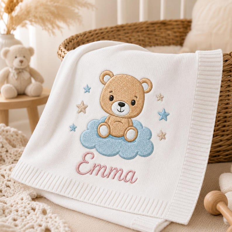 May include: A white knitted baby blanket featuring an embroidered design of a light brown teddy bear sitting on a blue cloud. The design includes small blue and beige stars and the name Emma embroidered in pink script lettering below the cloud.