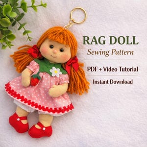 May include: A handmade rag doll keychain with orange yarn hair tied with red ribbons. The doll wears a pink polka-dot dress with a strawberry applique and red shoes. The text "RAG DOLL Sewing Pattern" is visible.