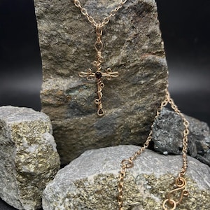 May include: A gold-colored wire-wrapped cross pendant necklace with a small, dark red gemstone at the center. The cross is displayed on a textured gray rock, with a gold chain and clasp visible.