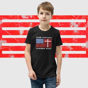 Könnte beinhalten: Schwarzes T-Shirt mit einer Grafik der amerikanischen Flagge, einem Kreuz und den Worten "ONE NATION UNDER GOD" in Weiß. Der Hintergrund hat ein rot-weißes Design.