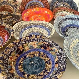 May include: A collection of handmade fabric bowls in various sizes and patterns. The bowls feature a spiral design with colorful fabrics, including floral, paisley, and abstract prints. The color palette includes blues, reds, oranges, and browns.