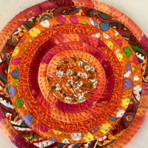 May include: Circular, handmade trivet with concentric rings of fabric in shades of orange, pink, and red. The center features a floral print, while other rings display polka dots and patterned fabrics. The trivet is stitched together with a contrasting thread.
