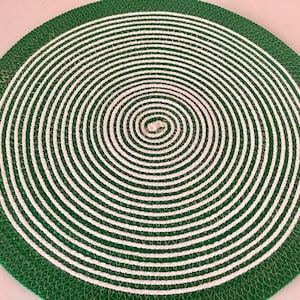 May include: Circular green and white woven placemat. The placemat features a spiral design with alternating bands of white and green. The outer edge is a solid green with a textured appearance. This decorative placemat is suitable for dining and kitchen decor.
