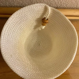 May include: A woven, off-white bowl with a spiral design. The bowl is round with a small, decorative wooden bead attached to the rim. The bowl is likely made of rope or a similar material.