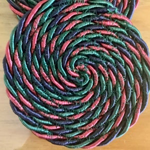 May include: Close-up of two spiral-shaped coasters. The coasters are made of tightly woven, colorful rope. The colors include shades of red, green, and blue. The coasters are stitched together with a dark thread.