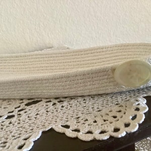 May include: A white woven basket with a large, oval, light-colored button closure. The basket is resting on a white, lacy, crocheted doily. The basket is made of a thick, woven material.