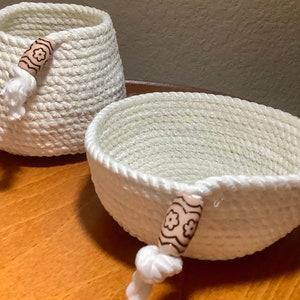 May include: Two handmade white rope baskets. One is a cylindrical shape, and the other is a shallow bowl shape. Both have decorative wooden beads with a floral design and a white tassel. The baskets are on a wooden surface.